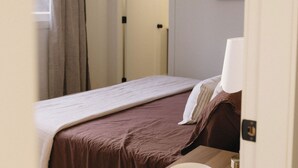 Ein Schlafzimmer mit einem Bett, einem Nachttisch mit Lampe, einem gerahmten Bild an der Wand und einer Tür zu einem anderen Raum.