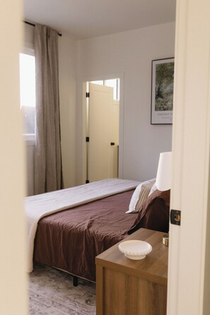 A bedroom with a bed, a bedside table with a lamp, a framed picture on the wall, and a door leading to another room. - The Vella | Private Modern Carriage House Near Shuswap Lake (Tappen)