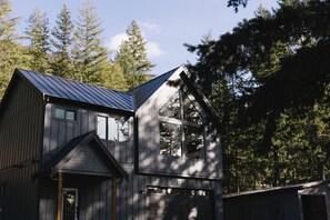 A two-story wooden house with a metal roof, surrounded by tall trees. - The Vella | Private Modern Carriage House Near Shuswap Lake (Tappen)