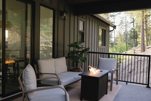 A covered outdoor seating area with a sofa and chairs, a table with a fire, and a view of a forested area. - The Vella | Private Modern Carriage House Near Shuswap Lake (Tappen)