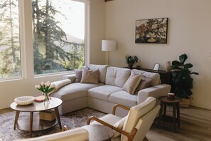 A living room with a sectional sofa, a small round table, a lamp, a painting, and a large plant. - The Vella | Private Modern Carriage House Near Shuswap Lake (Tappen)