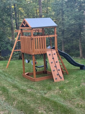 A wooden play structure with a slide, ladder, and climbing wall in a grassy area.