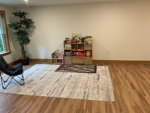 Una habitación con piso de madera, una silla negra, una planta grande en maceta, una estantería con juguetes y un tapete estampado.