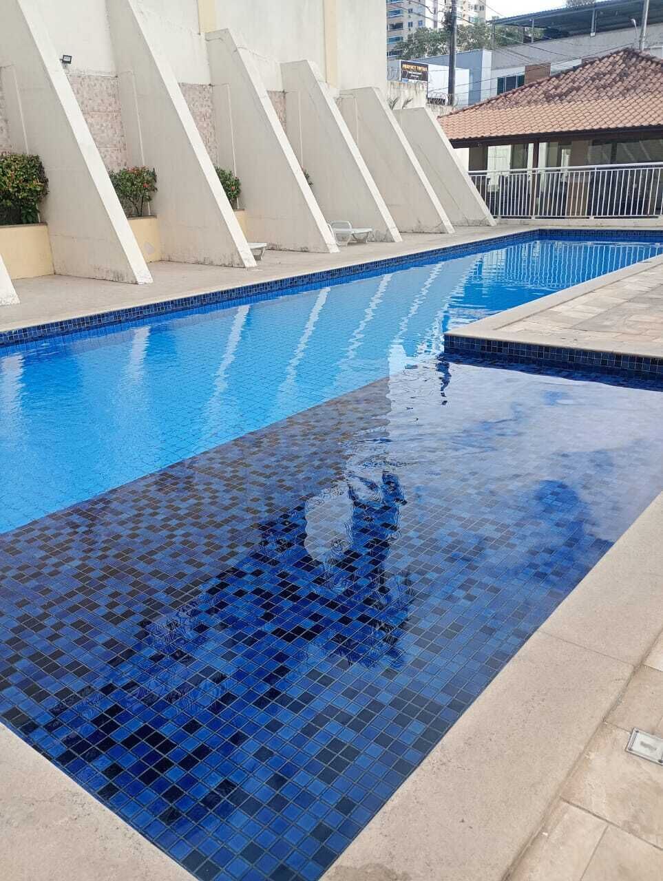 A swimming pool with blue tiles, a concrete deck, and a building with a tiled roof in the background.