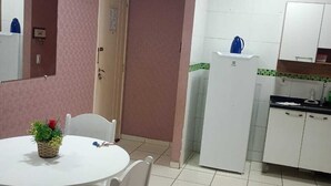 A compact kitchen with white cabinets, a refrigerator, and a sink. There is a dining area with a white table and chairs, and a small potted plant on the table. - Well-located apartment near beaches, shopping, etc. (Vila Velha)