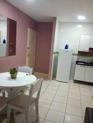 Eine kompakte Küche mit weißen Schränken, einem Kühlschrank und einer Spüle. Es gibt einen Essbereich mit einem weißen Tisch und Stühlen, und eine kleine Topfpflanze steht auf dem Tisch.