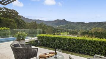 Une terrasse meublée de mobilier en osier, une table avec une bouteille et des verres, un plateau avec de la nourriture, et une vue sur des collines.