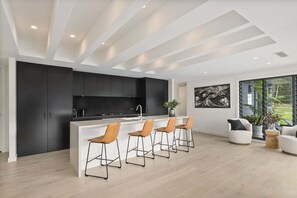 A modern kitchen with dark cabinets, a white island with bar stools, and a large window offering a view of greenery.
