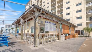 Un ristorante sulla spiaggia con posti a sedere all'aperto, un'insegna con il menu e vista su un edificio residenziale sullo sfondo.