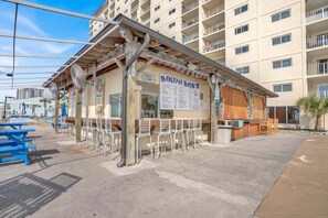 A beachfront restaurant with outdoor seating, a menu board, and a view of a residential building in the background. - Fall Deals + Snowbird Rates | Gulf Front Beach Escape (Panama City)
