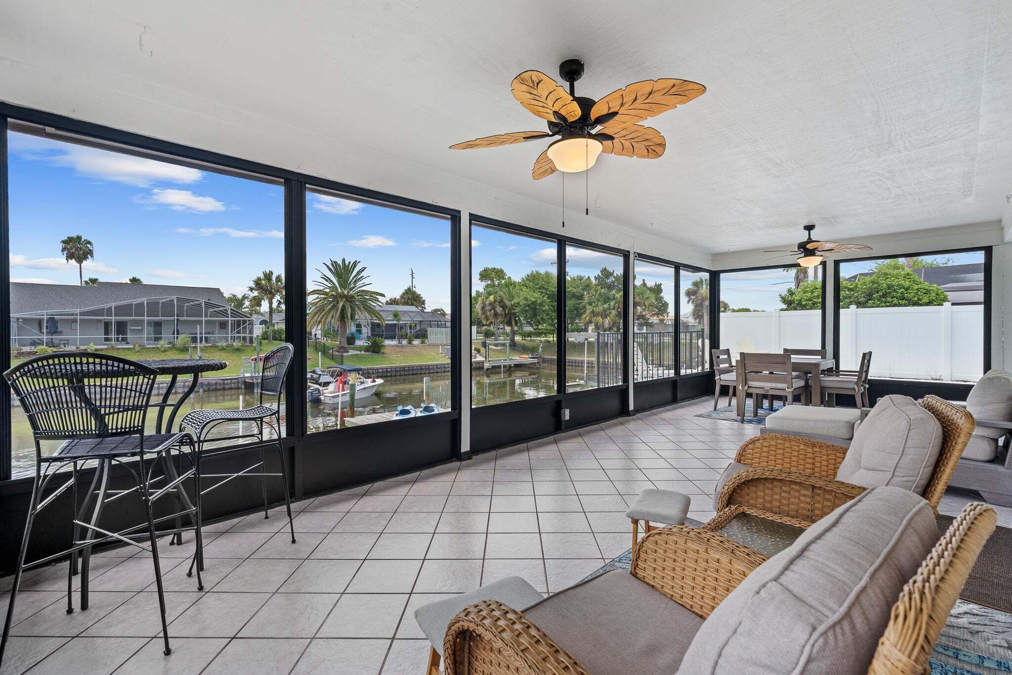 A spacious balcony with a view of a water body and palm trees, furnished with wicker sofas and metal chairs.
