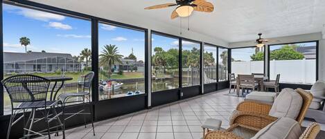 A spacious balcony with a view of a water body and palm trees, furnished with wicker sofas and metal chairs.