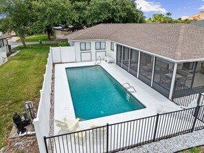 A rectangular swimming pool with a white deck, surrounded by a white fence, and a covered patio area with a view of the backyard. - Fall Deals + Snowbird Rates | Coastal Stay With Water Views (Palm Coast)