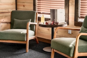 A cozy cabin interior with two green upholstered chairs, a small wooden table with a board game, and a window with blinds. - Golf Simulator/Rooftop Deck/Hot Tub/3BR/Sleeps 8! (Pigeon Forge)