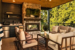 An outdoor kitchen with a stone fireplace, a dining area with a sofa and chairs, and a view of trees. - Golf Simulator/Rooftop Deck/Hot Tub/3BR/Sleeps 8! (Pigeon Forge)