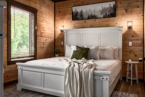 Een slaapkamer met een wit bed, houten wanden, een raam met zonwering, een schilderij boven het bed en een klein tafeltje met een lamp.