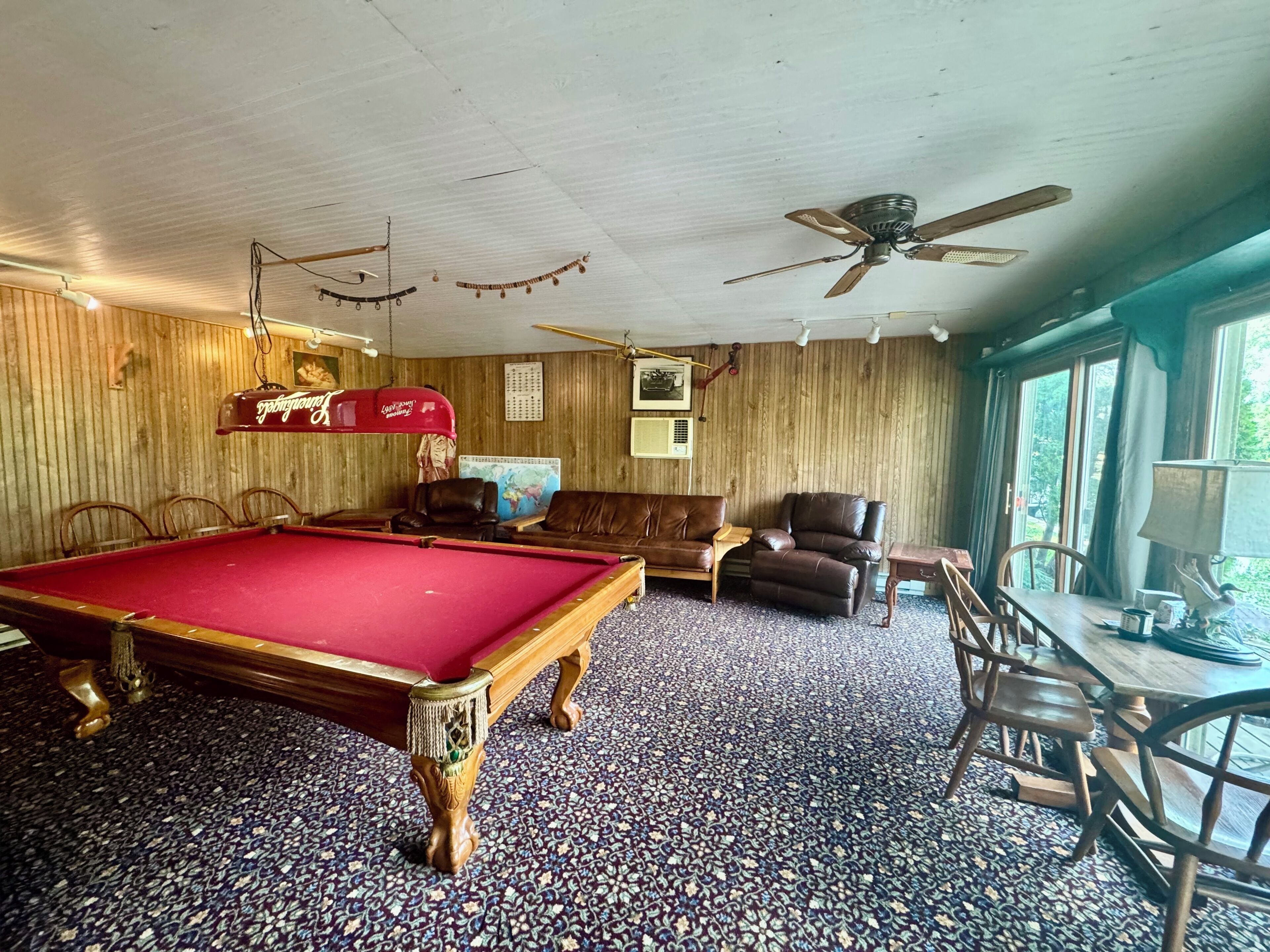 Une table de billard dans une pièce aux murs en bois, avec un ventilateur de plafond et une fenêtre donnant sur un beau paysage.