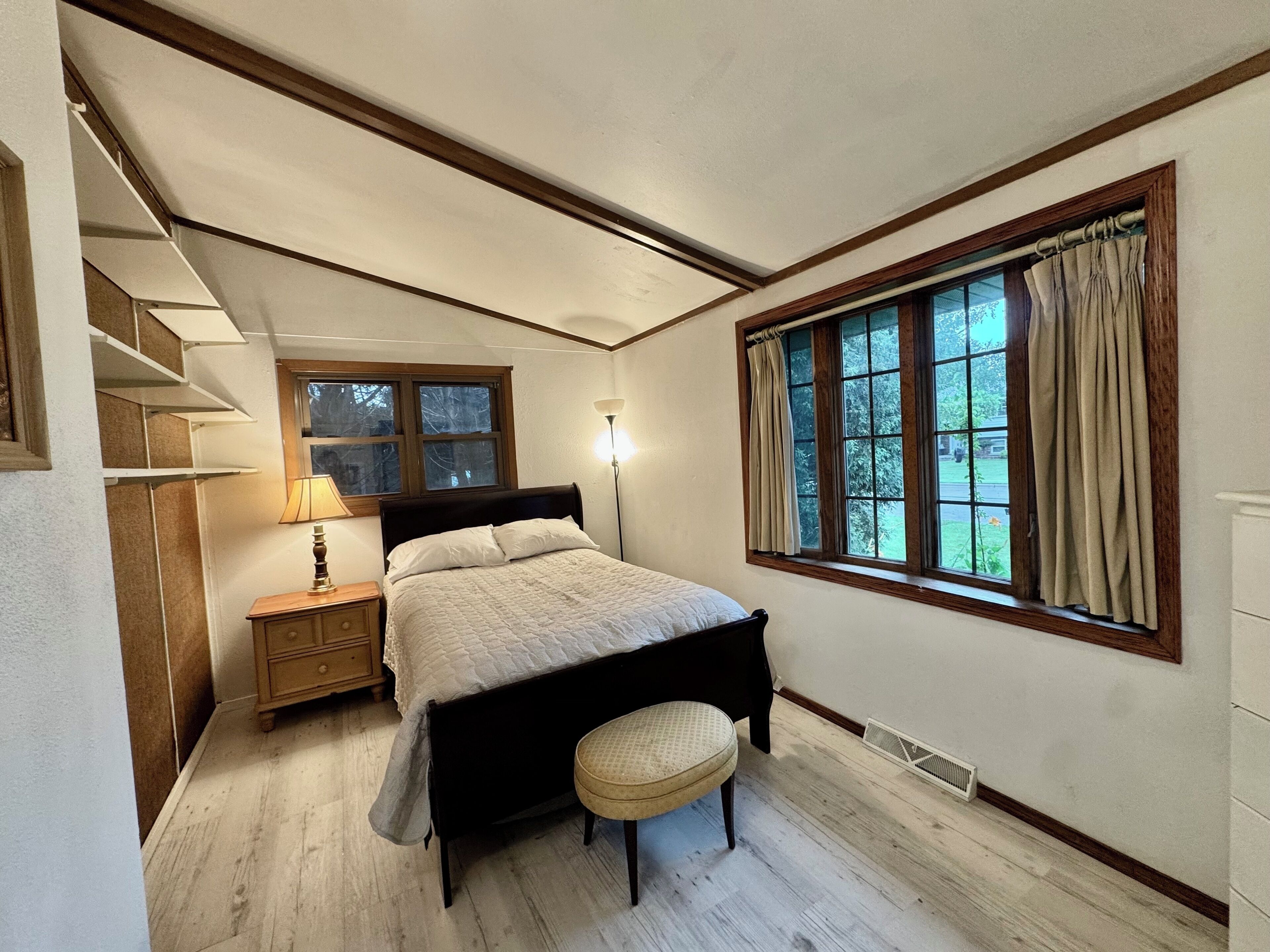 Ein Schlafzimmer mit Bett, Nachttisch, Lampe, Fenster mit Vorhängen und Blick ins Freie.
