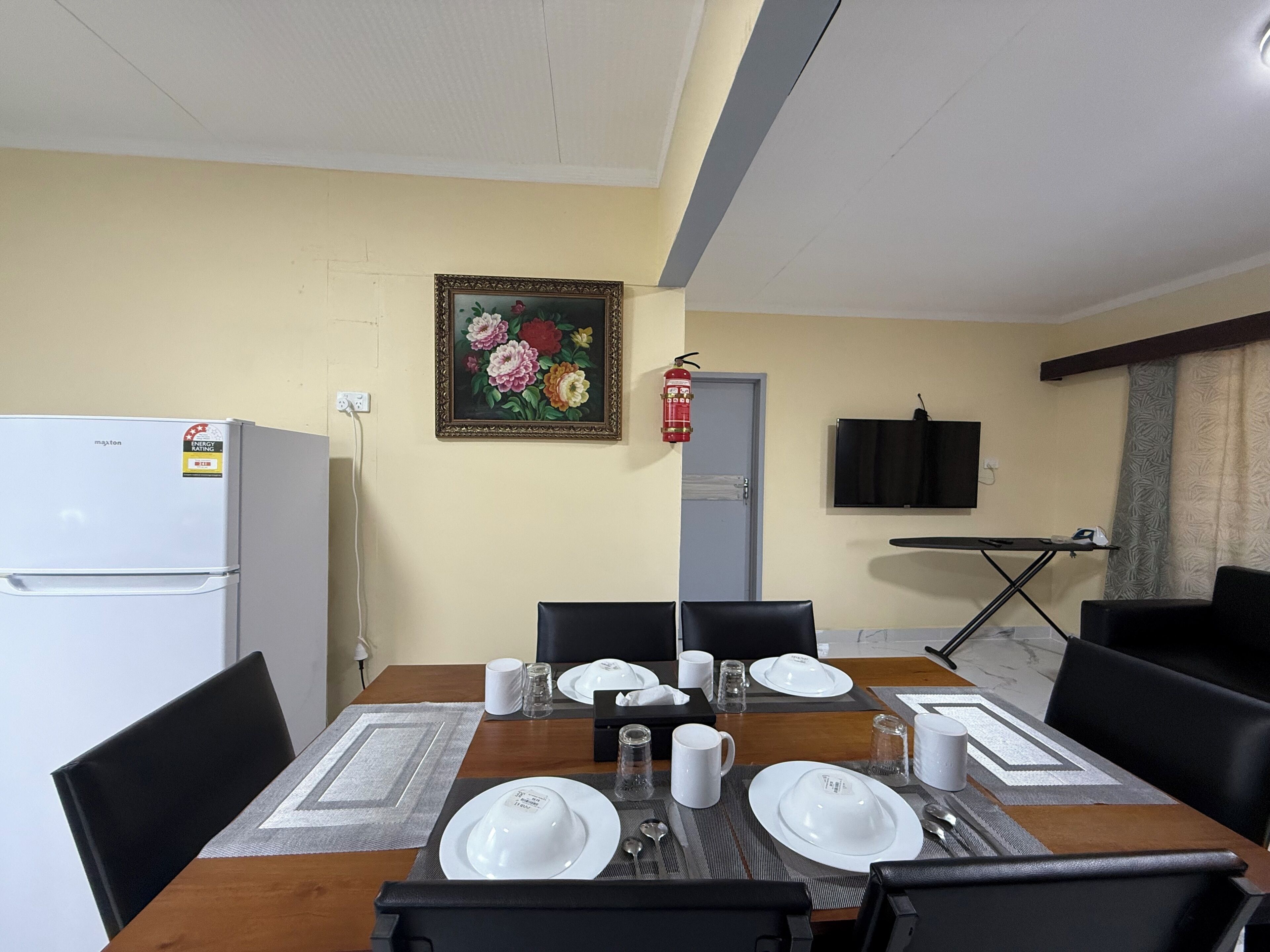 A dining area with a table set for four, a refrigerator, a fire extinguisher, and a television mounted on the wall.