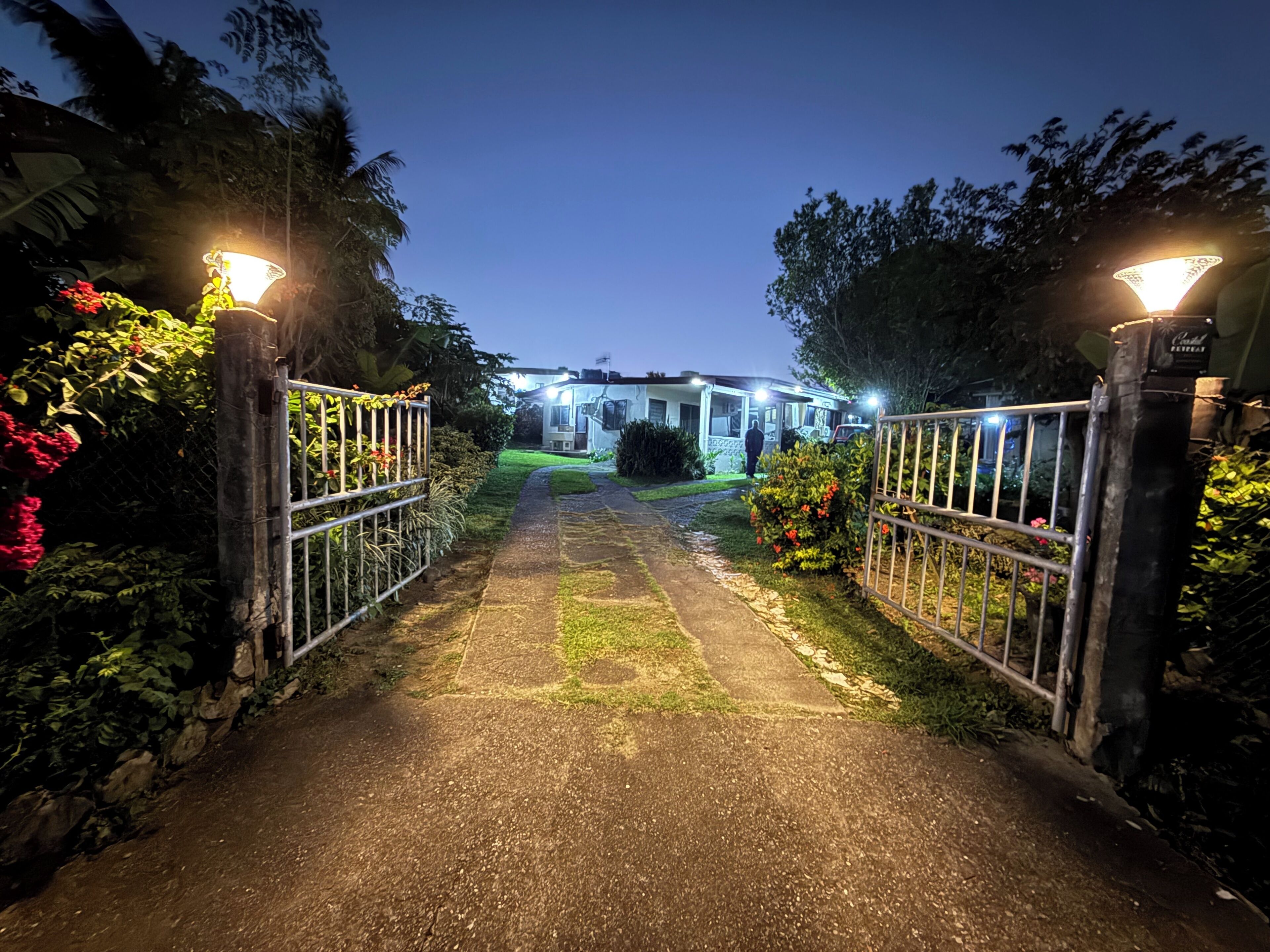 A gated pathway leading to a building with multiple lights, surrounded by greenery and a clear sky.