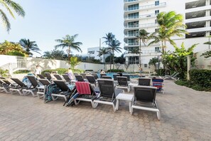 Uma área à beira da piscina com espreguiçadeiras, uma piscina e um edifício de vários andares ao fundo.