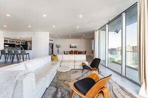 A modern living room with a white sofa, a dining area, and a large window offering a city view. - Oceanfront Penthouse with Pool and Beach Access (Hollywood)
