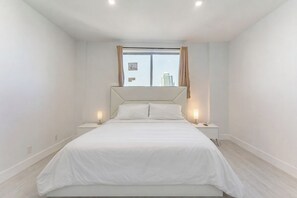 A bedroom with a bed, two bedside tables, a window with curtains, and a view of buildings. - Oceanfront Penthouse with Pool and Beach Access (Hollywood)