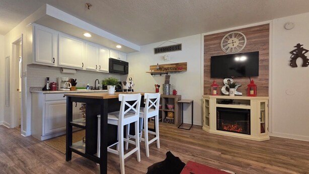 A kitchen with white cabinets, a microwave, and a bar area with white chairs. There's a fireplace with a TV above it and a clock on the wall. - Remodeled Westgate Cottage Near Pool & Rodeo Fun 316 (River Ranch)