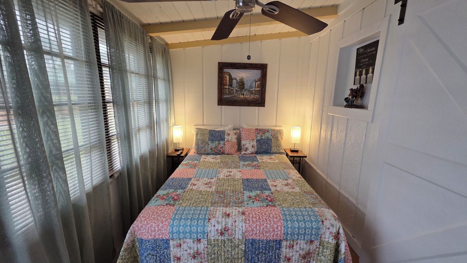 Une chambre à coucher avec un lit, deux lampes de chevet, un ventilateur de plafond et un tableau encadré au mur.