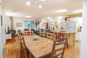 A modern kitchen with white cabinets, a central island, and a dining area with wooden chairs and a table. - Fernwood Cabin | Private Hot Tub + Wood-Burning Fire Near Mt Baker (Deming)