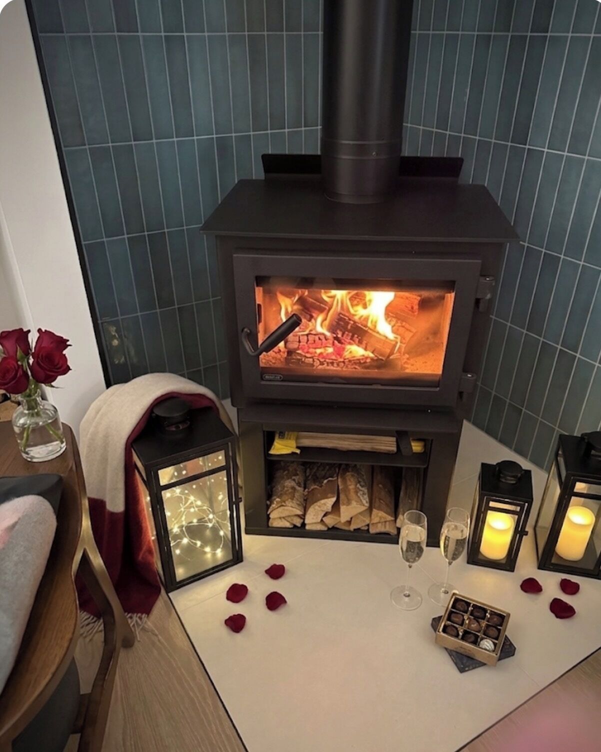 A wood-burning stove with a glass front, rose petals scattered on the floor, and two lit lanterns on a table.