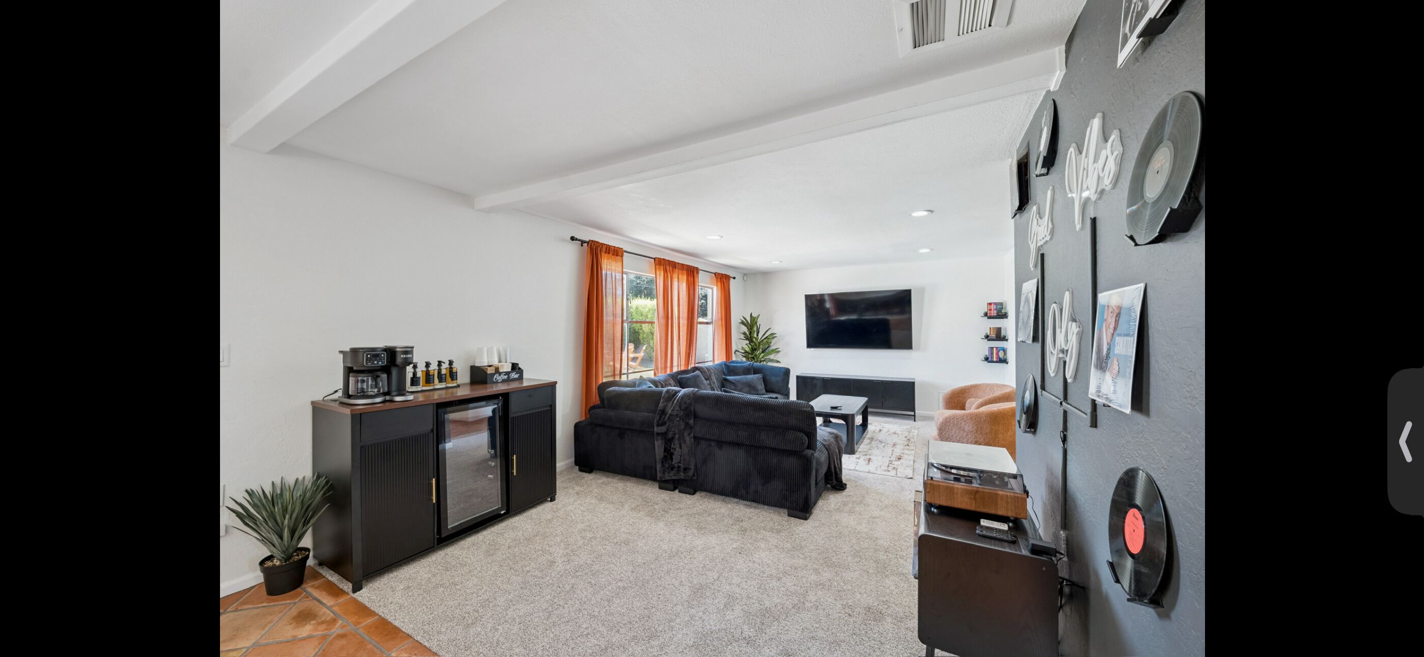Una sala de estar moderna con un sofá, una mesa de centro y un televisor de pantalla plana. Hay una cocina con un refrigerador y una cafetera.