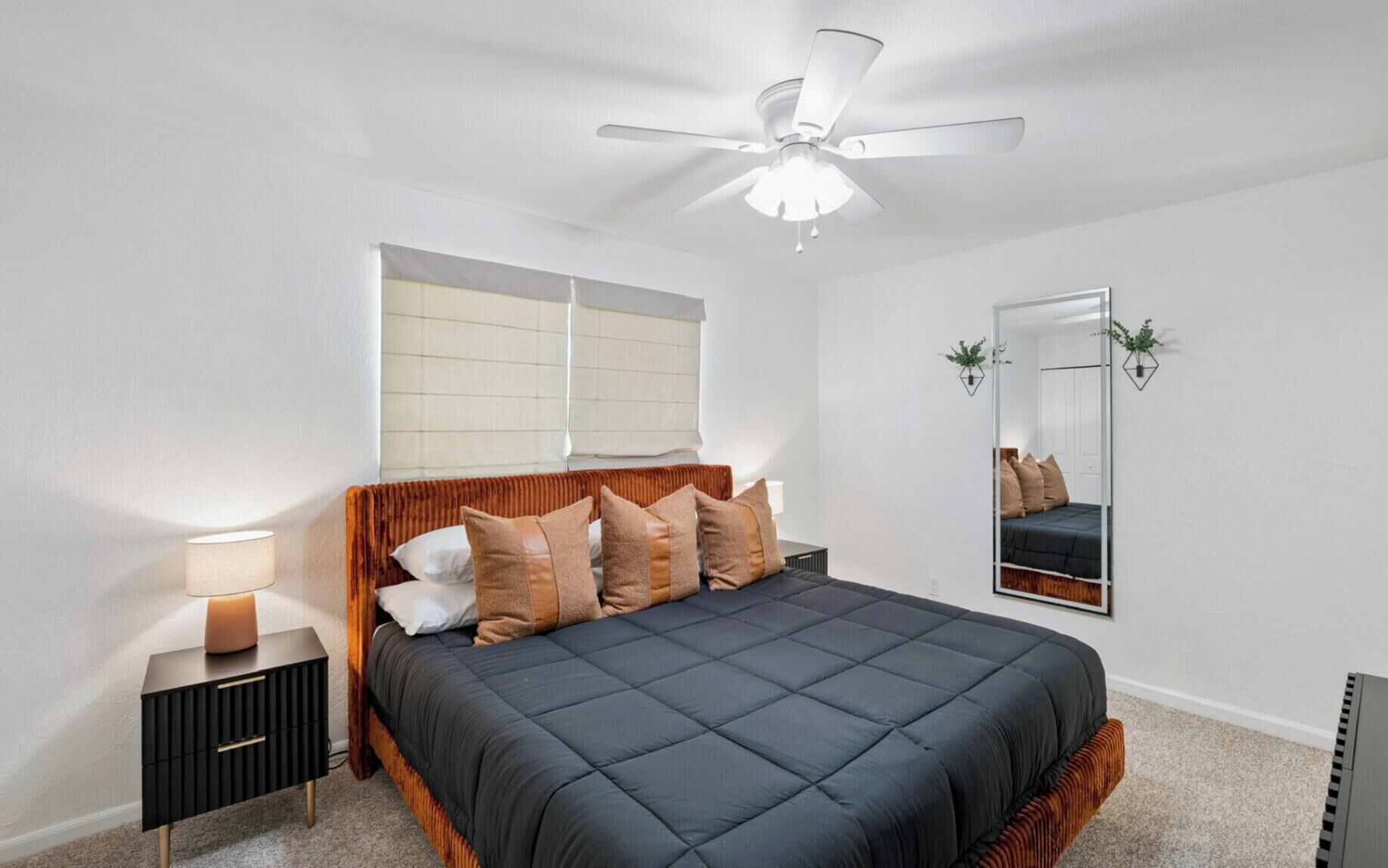 Un dormitorio con una cama, una mesita de noche, un ventilador de techo, una ventana con persianas, un espejo y una planta colgante en la pared.