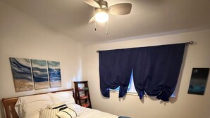 Een slaapkamer met een bed, een plafondventilator, een raam met gordijnen en kunst aan de muur.