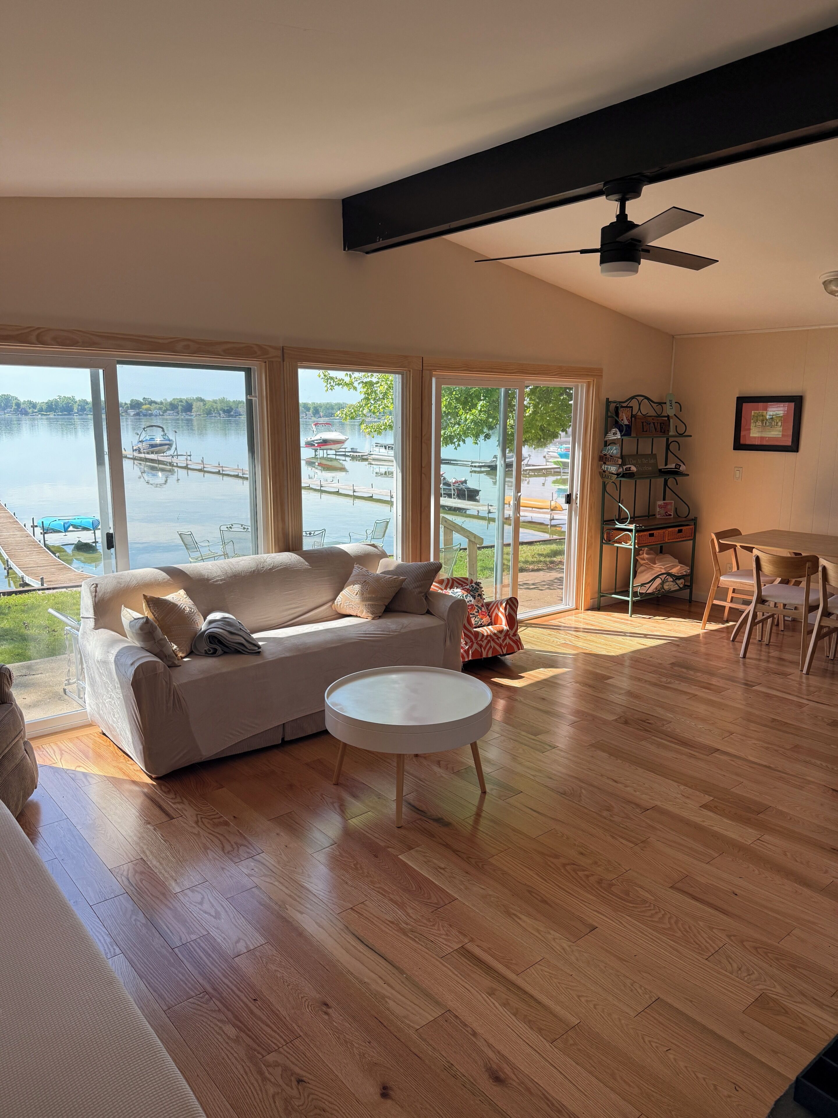 A spacious living room with a large window overlooking a lake, a wooden floor, a white sofa, and a dining area with chairs.