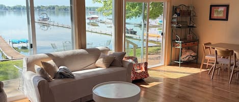 A spacious living room with a large window overlooking a lake, a wooden floor, a white sofa, and a dining area with chairs.