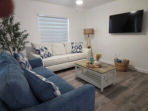 Ein Wohnzimmer mit einer Couch, einem Couchtisch, einem an der Wand montierten Fernseher und einem Fenster mit Jalousien.