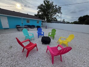 A patio area with colorful Adirondack chairs arranged in a circle, a building with a blue exterior, and a tree in the background. - 3 Minutes to Anna Maria Island. Boat, RV & Jet Ski Parking + Large Private Yard (Bradenton)