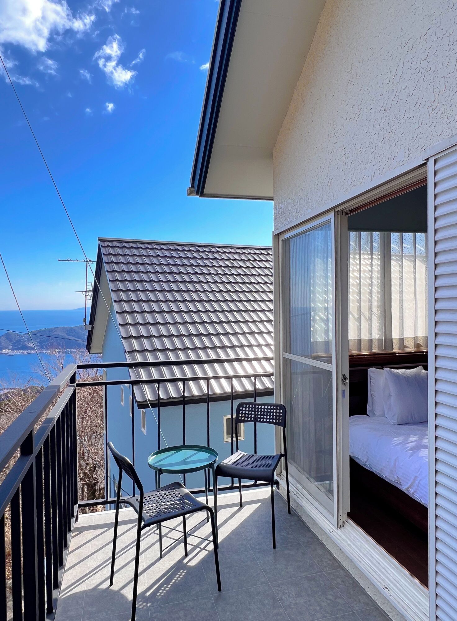 A balcony with a table and chairs, a bed with white bedding, and a view of the sea.