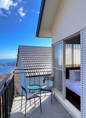 Un balcón con mesa y sillas, una cama con ropa de cama blanca, y vista al mar.