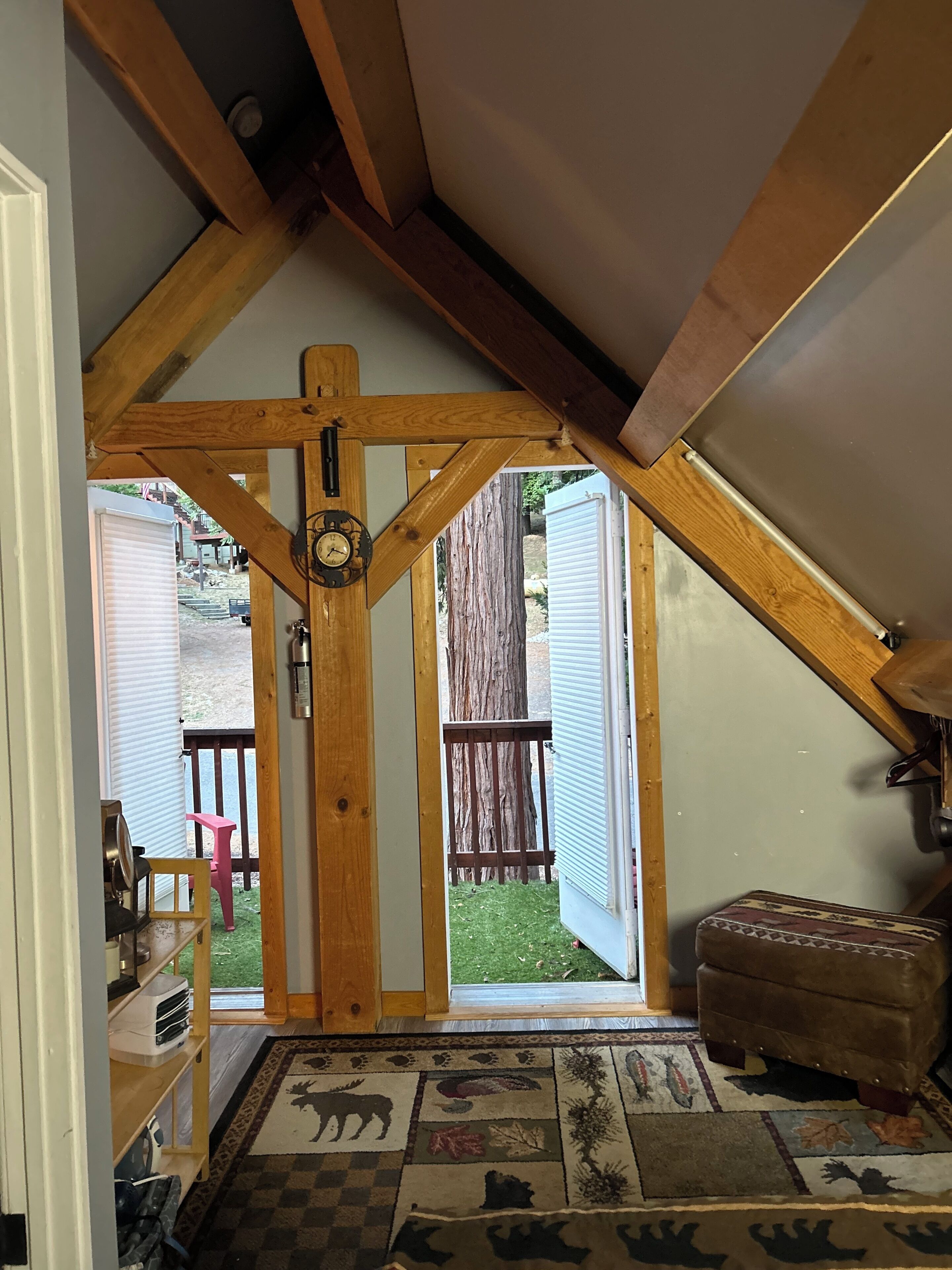 A room with a sloped ceiling, wooden beams, a clock, a patterned rug, and a view of a backyard with a wooden deck and a tree.