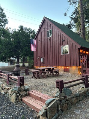 A rustic wooden building with a picnic table and benches, a flagpole with an American flag, and a stone wall.