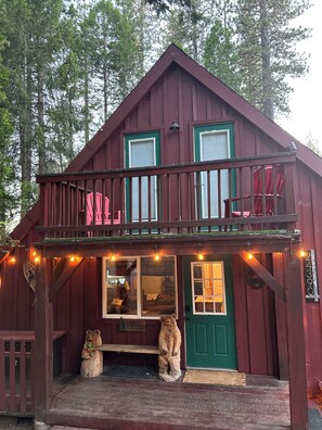 A two-story wooden cabin with a balcony, a green door, and a small front porch with a bear statue.
