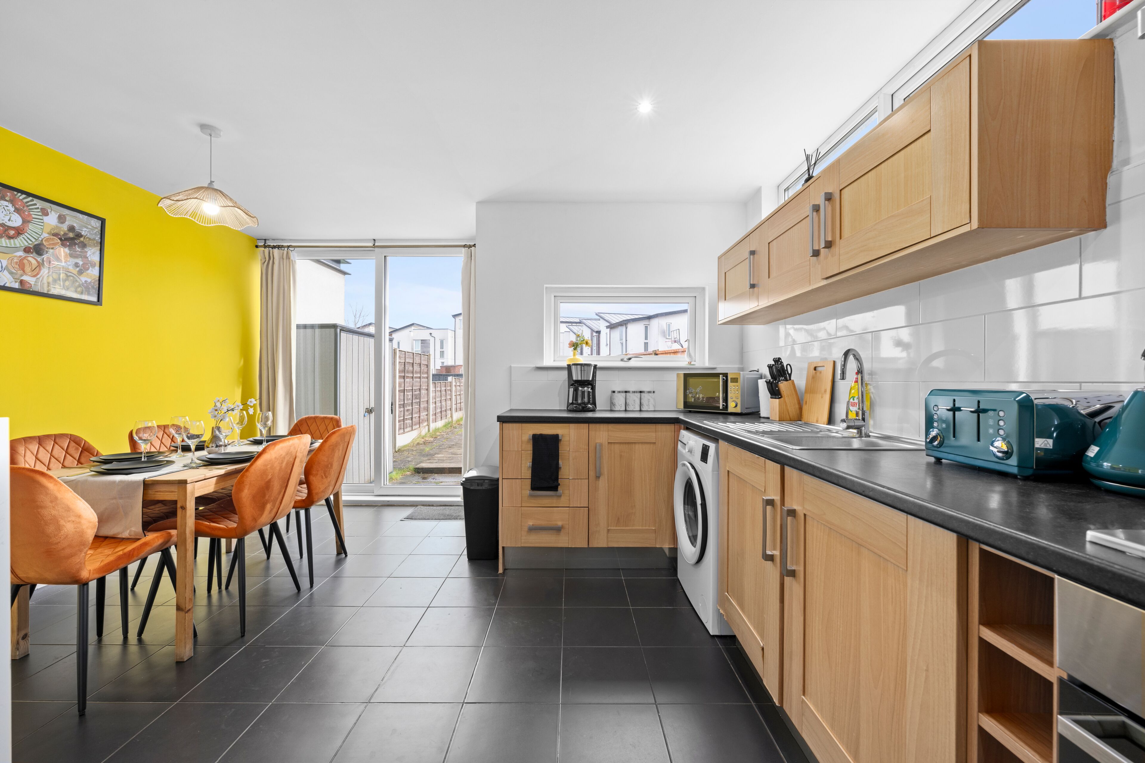 A modern kitchen with wooden cabinets, a washing machine, and a dining area with orange chairs.