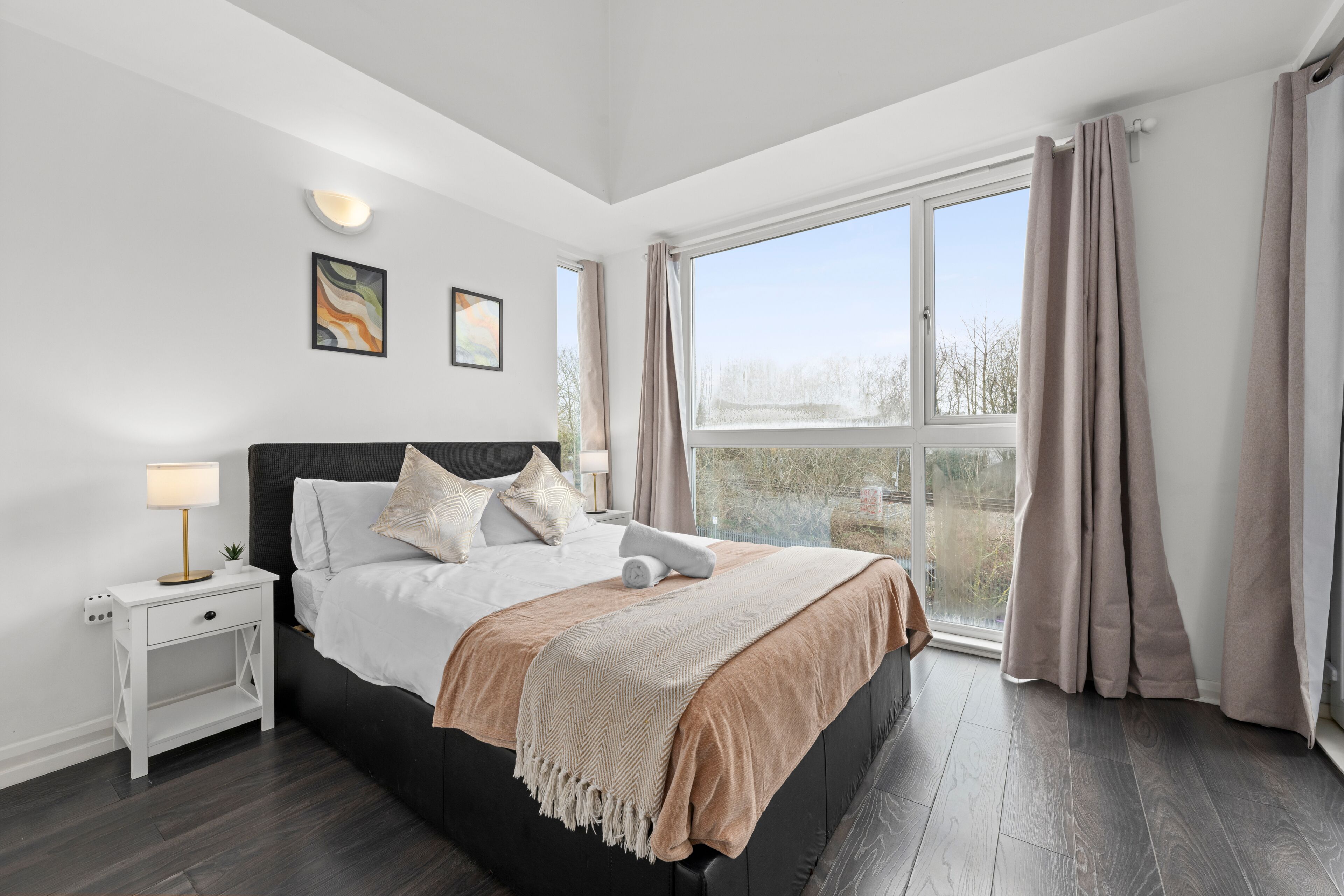 A modern bedroom with a large bed, bedside tables, a window with curtains, and wall-mounted lighting.