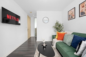 Een moderne woonkamer met een flatscreen televisie waarop 'NETFLIX' staat, een groene bank met kleurrijke kussens, een ronde spiegel en een potplant.