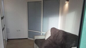 Ein Zimmer mit grauem Sofa, weißem Sessel, einem Fenster mit Jalousien und einem Fliesenboden.
