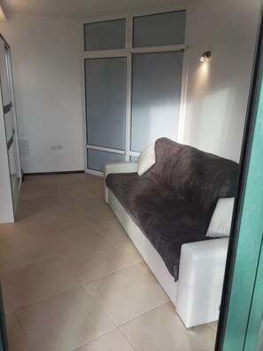 A room with a grey sofa, a white armchair, a window with blinds, and a tiled floor. - Apartment Sleeps 2 with Pool & Balcony (Balchik)
