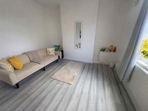 A living room with a beige sofa, a small rug, a white wall with a mirror, and a window with curtains. - 2-Bed House, Sleeps 4 with Parking & Garden (Bishop Auckland)