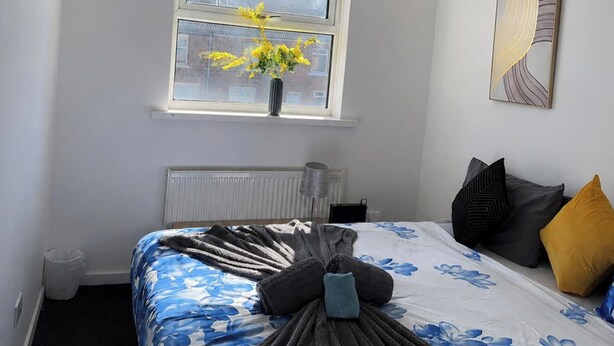 Een slaapkamer met een bed, een raam met bloemen, een radiator en een schilderij aan de muur.